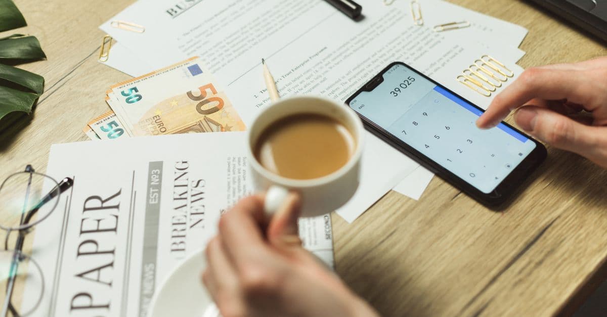 Schreibtisch mit Unterlagen, Kaffee, Geldscheinen und Smartphone mit Taschenrechner aus der Vogelperspektive