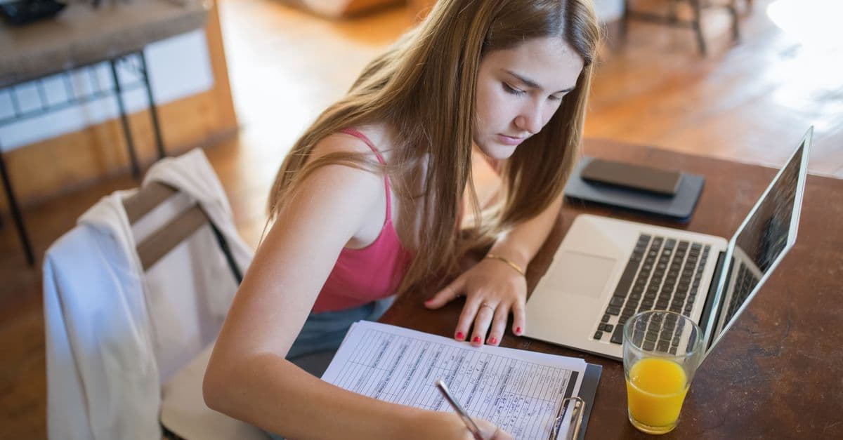 Fokussierte Frau arbeitet zu Hause am Laptop, daneben Klemmbrett und ein Glas Orangensaft.