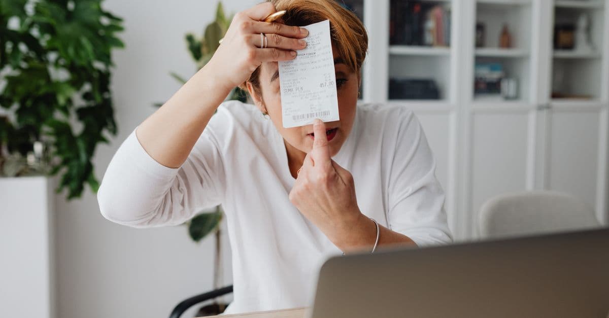 Frau im Homeoffice hält einen Kassenbon in die Kamera, Laptop und Smartphone auf dem Schreibtisch