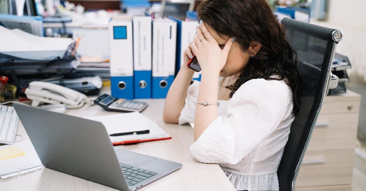 Gestresste Frau sitzt im Büro am Schreibtisch und telefoniert, den Blick angespannt auf den Tisch gerichtet.