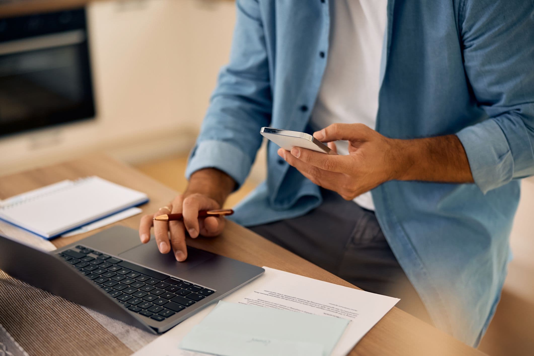 Mann am Schreibtisch nutzt Smartphone und Laptop, daneben Notizbuch und Unterlagen.