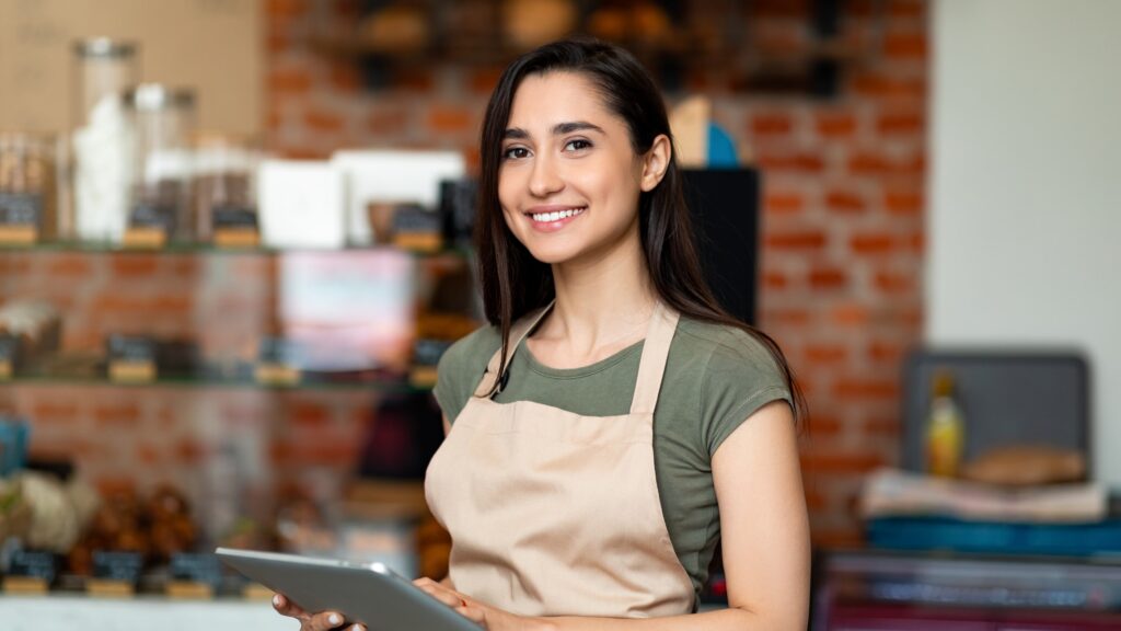 Junge Frau im Café mit Schürze und Tablet, arbeitet in einem Minijob im Service.
