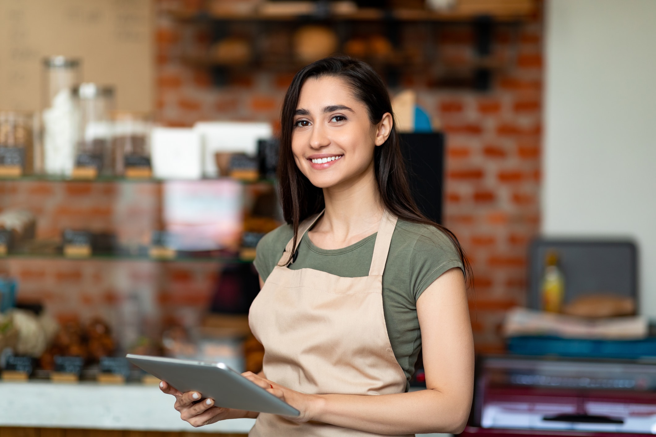 Junge Frau im Café mit Schürze und Tablet, arbeitet in einem Minijob im Service.
